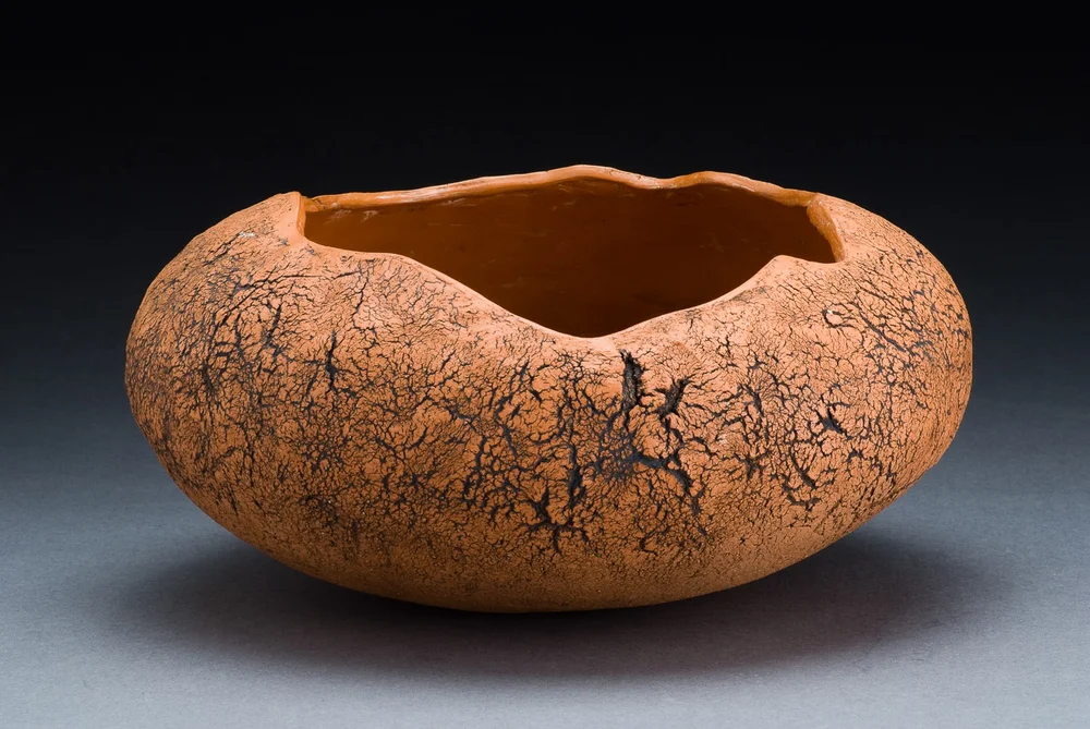 A rounded, organic-shaped ceramic bowl with a textured, cracked orange surface and dark crevices, photographed against a plain gray background